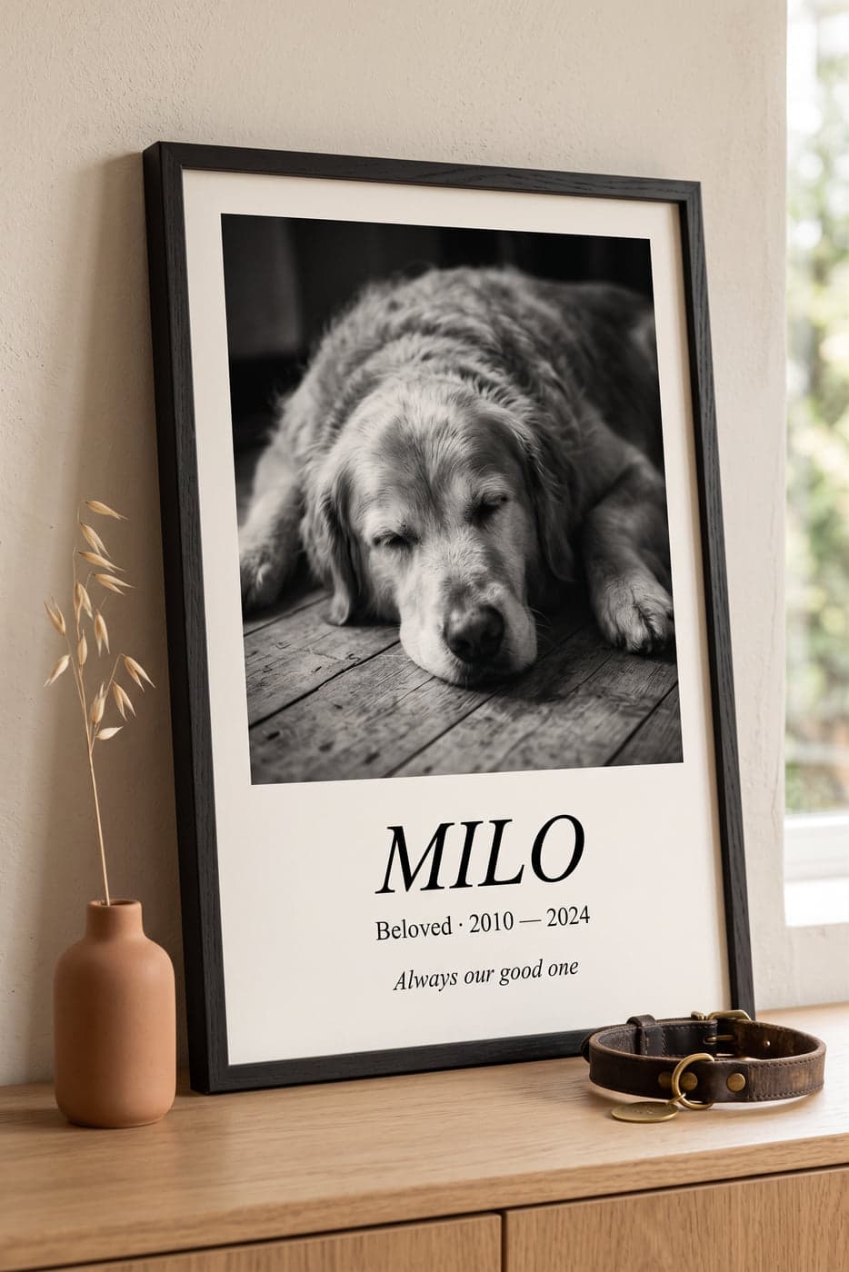 A framed black-oak memorial poster on an oak console table showing a black-and-white photo of a golden retriever, with the caption MILO · Beloved 2010–2024 · Always our good one